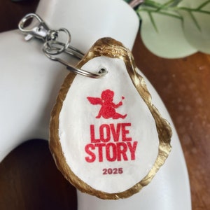 May include: A gold-painted oyster shell keychain with a red and white design. The design features a cupid with a heart and the words "LOVE STORY 2025".