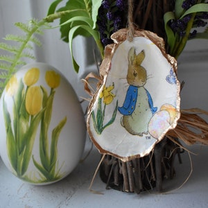 Może przedstawiać: Złota, ozdobna ozdoba w kształcie jajka wielkanocnego z ilustracją Królika Piotrusia. Jajko jest ozdobione niebieskim królikiem, motylem i żółtymi narcyzami. Ozdoba jest zawieszona na gałązce.