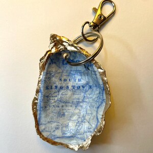 May include: A handmade keychain featuring a map of South Kingstown, Rhode Island, inside a gold-edged oyster shell. The shell is attached to a metal clasp and key ring. The map is printed in blue on a white background.