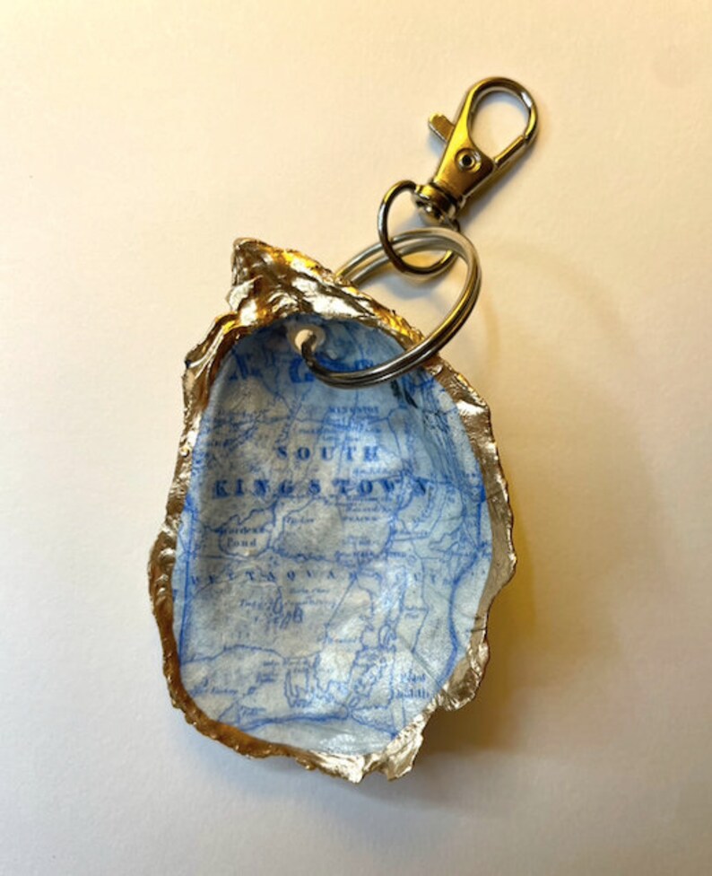 May include: A handmade keychain featuring a map of South Kingstown, Rhode Island, inside a gold-edged oyster shell. The shell is attached to a metal clasp and key ring. The map is printed in blue on a white background.