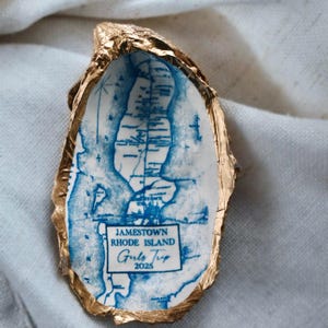May include: An oyster shell with a gold-painted edge, featuring a blue map of Jamestown, Rhode Island. The text "Jamestown Rhode Island Girls Trip 2025" is printed on the shell, making it a unique souvenir or gift.