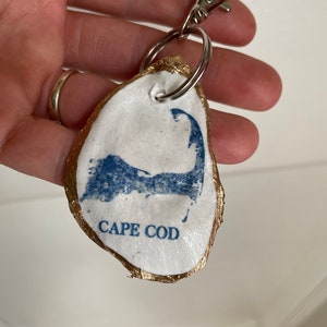 May include: A handmade oyster shell keychain with a gold-painted edge. The white shell features a blue Cape Cod outline and text. Includes a silver-colored clasp and keyring.