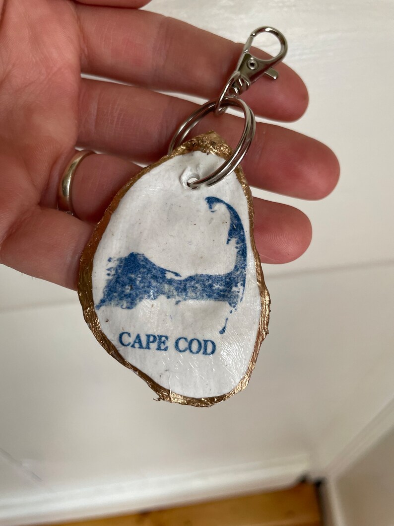 May include: A handmade oyster shell keychain with a gold-painted edge. The white shell features a blue Cape Cod outline and text. Includes a silver-colored clasp and keyring.