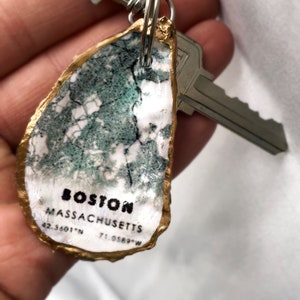 May include: A unique keychain featuring a painted oyster shell with a map design. The shell is edged in gold and displays the text "BOSTON MASSACHUSETTS" along with coordinates. A silver key and clasp are attached.