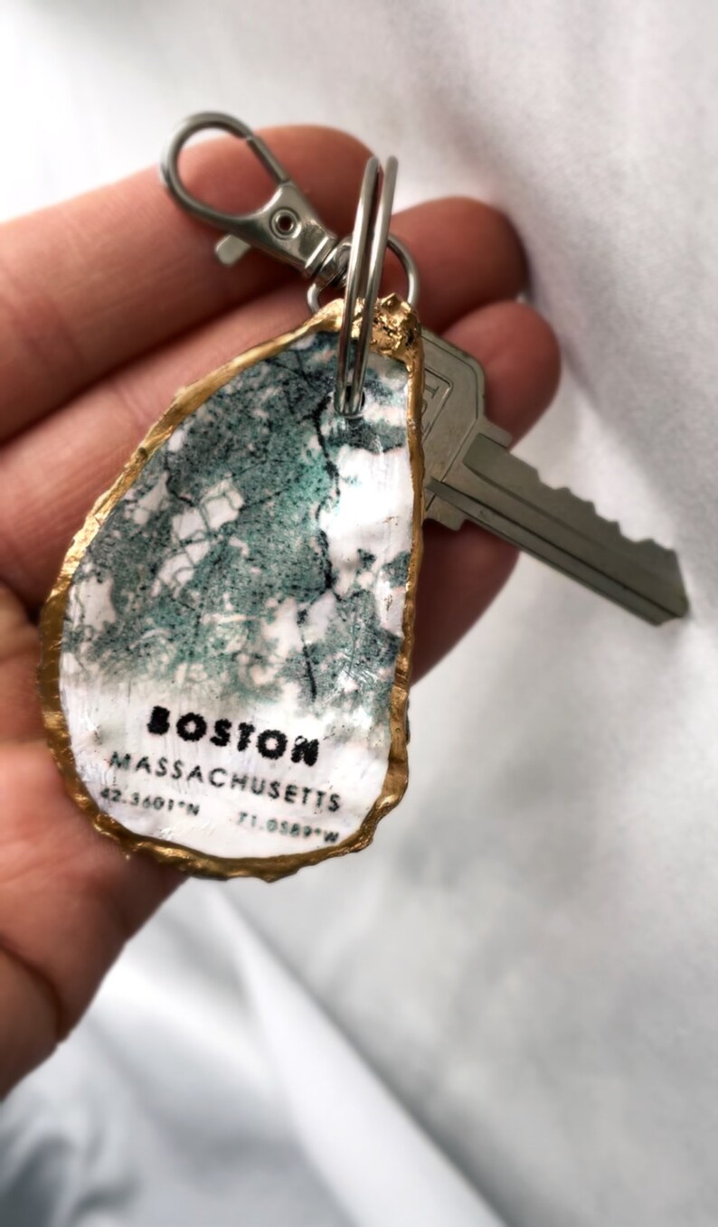 May include: A unique keychain featuring a painted oyster shell with a map design. The shell is edged in gold and displays the text "BOSTON MASSACHUSETTS" along with coordinates. A silver key and clasp are attached.
