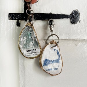 May include: Two oyster shell keychains. One features a map of Boston, Massachusetts, with the city name and coordinates. The other displays a blue Cape Cod outline with the text "CAPE COD". Both have gold-painted edges and silver key rings.