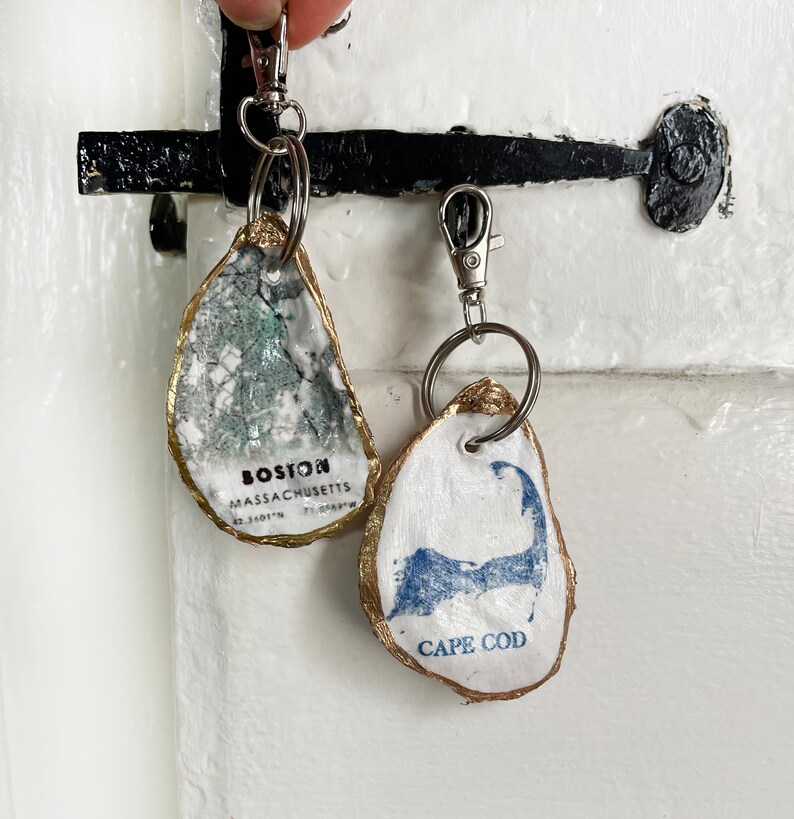 May include: Two oyster shell keychains. One features a map of Boston, Massachusetts, with the city name and coordinates. The other displays a blue Cape Cod outline with the text "CAPE COD". Both have gold-painted edges and silver key rings.