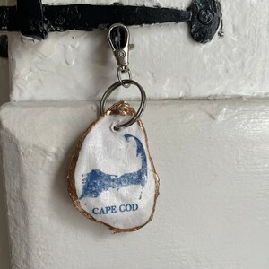 May include: A white oyster shell keychain with a blue Cape Cod map design and gold trim. The keychain is attached to a silver clasp and key ring. The text "CAPE COD" is printed below the map.