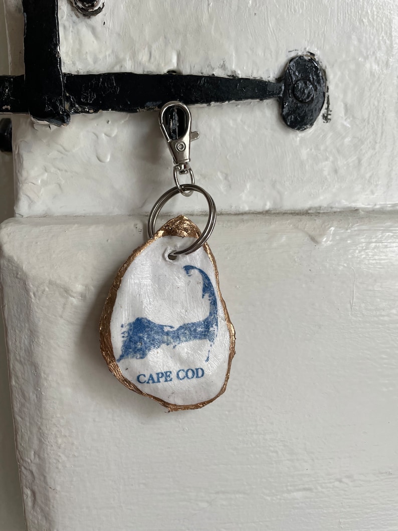 May include: A white oyster shell keychain with a blue Cape Cod map design and gold trim. The keychain is attached to a silver clasp and key ring. The text "CAPE COD" is printed below the map.