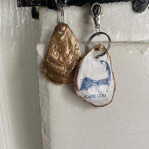 May include: Two oyster shell keychains. One is painted gold, the other is white with a blue Cape Cod design and the words "Cape Cod". Both have silver key rings and clasps.