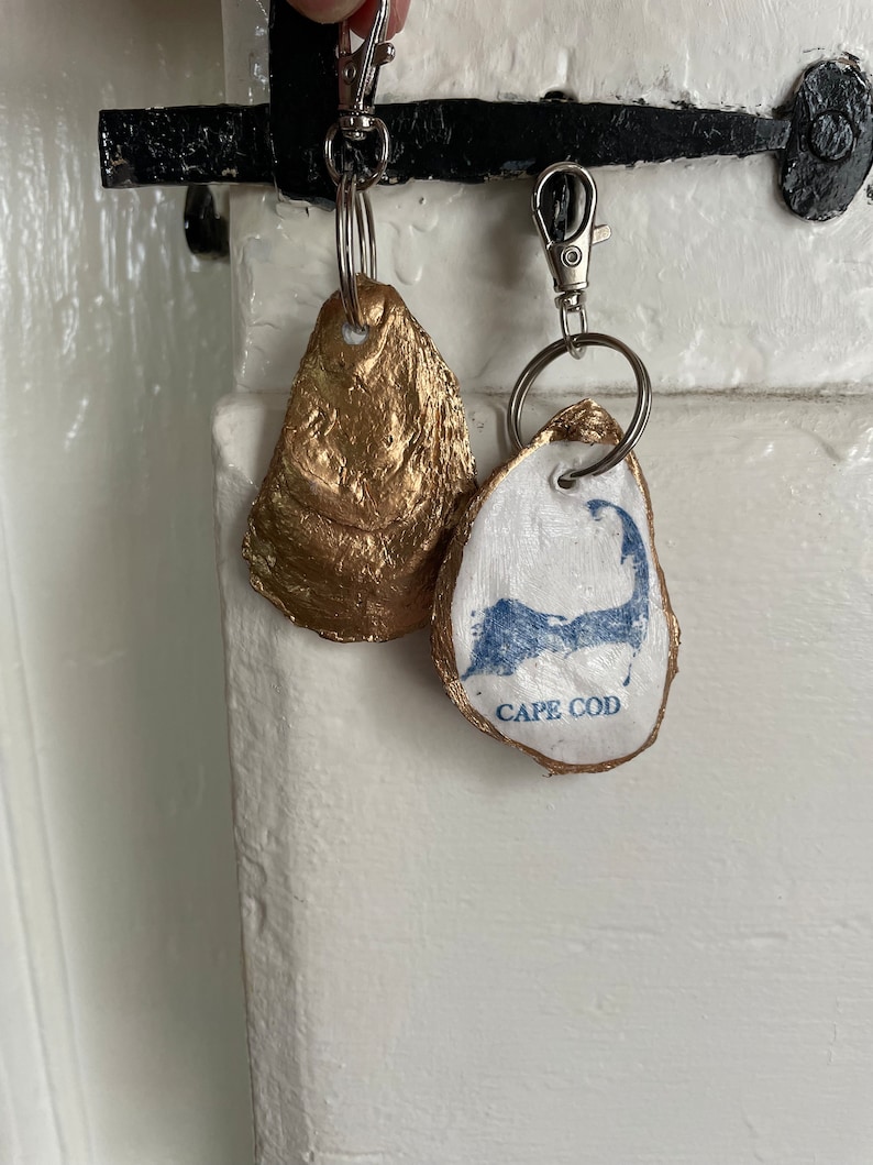 May include: Two oyster shell keychains. One is painted gold, the other is white with a blue Cape Cod design and the words "Cape Cod". Both have silver key rings and clasps.