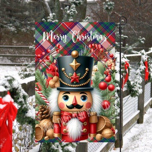 Puede incluir: Una bandera de jardín navideña con un diseño de cascanueces. La bandera tiene un fondo a cuadros con las palabras "Merry Christmas". El cascanueces lleva un sombrero negro y un uniforme rojo, rodeado de decoraciones festivas.