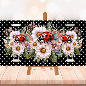 May include: A license plate with a black background and white polka dots. The plate features a floral arrangement of white and pink flowers with three red ladybugs. The license plate is displayed on a wooden easel.