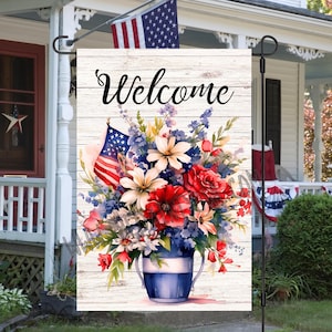 May include: A welcome garden flag with a whitewashed wood background. The flag features a blue and white vase with red, white, and blue flowers. A small American flag is tucked into the bouquet. The text "Welcome" is written in black cursive.