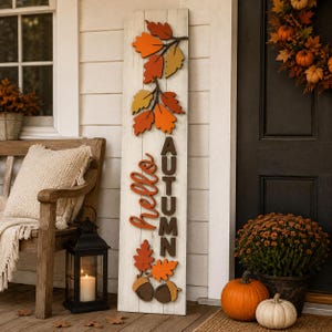 Pode incluir: Uma placa de madeira branca, alta e retangular, com o texto "hello AUTUMN" em letras em relevo castanho-escuras e cor de laranja. A placa está decorada com recortes de madeira tridimensionais em camadas de folhas de outono e bolotas.