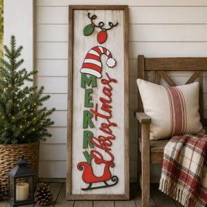Puede incluir: Un letrero de madera rectangular alto con un acabado blanco desgastado y un marco de madera oscura. El letrero presenta un gorro a rayas rojas y blancas, el texto verde MERRY, el texto cursivo rojo Christmas y un trineo rojo con luces.