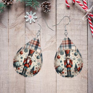 May include: A pair of dangle earrings with a red and white plaid pattern and a round image of a nutcracker in a red and blue uniform. The nutcracker is holding a sword and has a mustache. The earrings are on a wooden background.