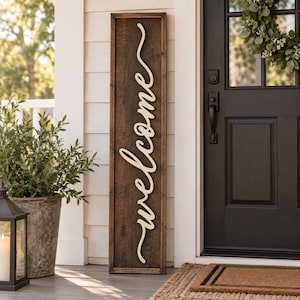 May include: A tall, rectangular wooden sign with a dark brown stained finish. The word welcome is written vertically in a light-colored, cursive script font, featuring a raised, three-dimensional design centered on the wooden board.