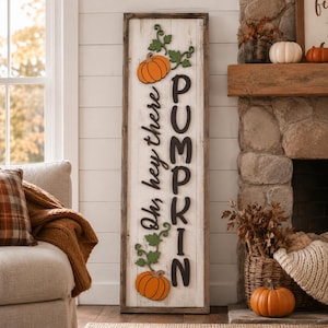 May include: A tall, rectangular wooden sign with a white-washed finish and a dark wood frame. The sign features the black text "Oh, hey there PUMPKIN" vertically, accented by two orange pumpkins with green vines at the top and bottom of the board.