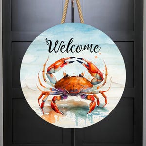 May include: A round wooden door hanger with a watercolor painting of a crab on a beach background. The words "Welcome" are written in black script above the crab.