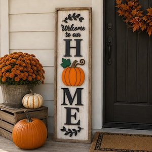 May include: A tall, rectangular wooden sign with a white distressed finish and a dark wood frame. The sign features the black text Welcome to our, followed by the word HOME, where the letter O is replaced by an orange pumpkin with a green leaf.