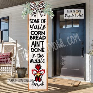Könnte beinhalten: Ein vertikales Veranda-Schild aus Holz mit weißer Holzmaserung, verziert mit einer schwarzen Schleife mit Kuhmuster und grünem Blattwerk. Der Text lautet: SOME OF Y'ALLS CORN BREAD AIN'T DONE IN THE MIDDLE, über einem Cartoon-Huhn mit einer roten Brille.