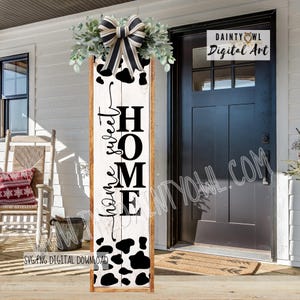Könnte beinhalten: Ein Holzschild mit den Worten "sweet home" in schwarzer Schrift. Das Schild hat einen weißen Hintergrund mit schwarzen Kuhflecken und ist mit Grün und einer schwarz-weiß gestreiften Schleife versehen. Das Schild lehnt an einem weißen Haus.