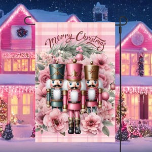 Puede incluir: Una bandera de jardín de temática navideña rosa y blanca con el texto "Merry Christmas" y tres figuras de cascanueces con detalles en rosa, verde y dorado.