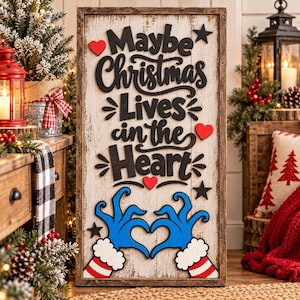 May include: A rectangular wooden sign with a distressed white finish and a dark wood frame. The sign features black 3D lettering that reads "Maybe Christmas Lives in the Heart," accented by red hearts, black stars, and blue cartoon hands forming a heart.