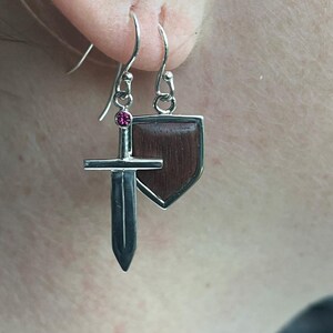 May include: Silver earrings with a small sword charm and a wooden shield charm. The sword charm has a pink gemstone.
