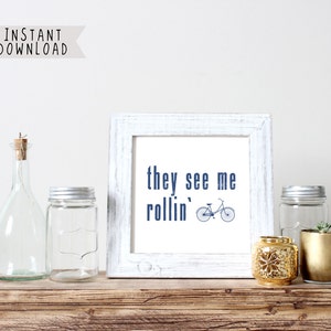May include: A framed print with the text "they see me rollin'" and a bicycle illustration. The print is in a white distressed frame, displayed on a wooden surface with glass jars, a gold candle holder, and a small potted plant.