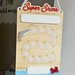 May include: Wooden "Super Shark" little wins tracker with a red title and a shark graphic. Includes spaces for tracking wins, a reward section, and a crab accent. Features a white ribbon for hanging.