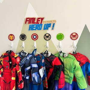 May include: A wooden wall sign reading "Finley IT'S TIME TO HERO UP!" mounted above six circular superhero icons. Below the icons, six white adhesive hooks hold various superhero costumes, including Iron Man, Black Panther, and Spider-Man.