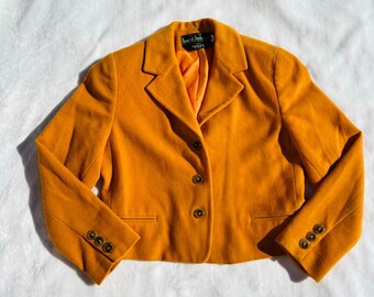 Könnte beinhalten: Ein senfgelber Blazer mit Reverskragen und drei goldenen Knöpfen. Der Blazer hat eine verkürzte Länge und ist aus einem weichen, strukturierten Stoff gefertigt.