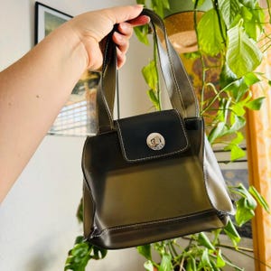 May include: A black Guess handbag with a rectangular shape and a flap closure. The bag features white stitching and a silver-toned Guess logo on the flap. The handles are a darker shade of gray.