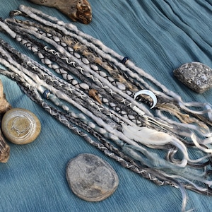May include: A set of grey, white, and black dreadlocks with decorative beads, feathers, and a crescent moon charm. The dreadlocks are displayed on blue fabric with stones and driftwood.