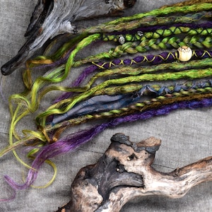 May include: Close-up of colourful dreadlocks in shades of green, purple, and yellow, with decorative beads and accents. The dreadlocks are arranged on a grey surface with pieces of driftwood.