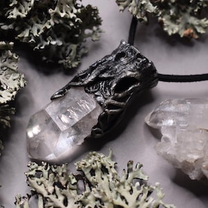 Op de afbeelding: Een zilveren ketting met een hanger met een helder kwartskristal dat is ingekapseld in een gedetailleerd, boomvormig ontwerp. De hanger hangt aan een zwart koord en is omgeven door grijs mos.