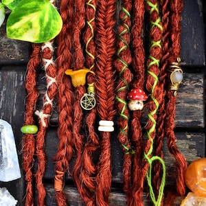 May include: Close-up of red dreadlocks adorned with decorative elements. The dreads are styled with green and white thread, and feature charms like a mushroom, a pentagram, and a gemstone. The overall aesthetic is bohemian and artistic.