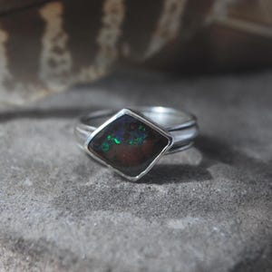 Peut inclure: Une bague en argent ornée d'une pierre sombre en forme de cerf-volant avec des éclats irisés verts et bleus. L'anneau est lisse et métallique, maintenant la pierre dans un sertissage simple. La pierre présente une base brun foncé avec des reflets.