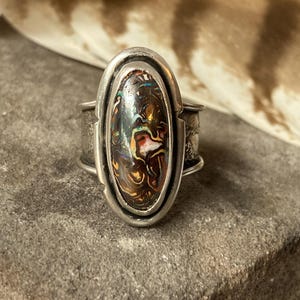 Op de afbeelding: Een ring van sterling zilver met een grote, ovale, donkere steen met wervelende insluitingen van bruin, oranje en iriserend blauw. De metalen band heeft een brede, getextureerde afwerking met een gladde zetting rondom de centrale steen.