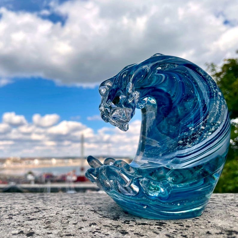 Pode incluir: Escultura em vidro artesanal de uma onda, mostrando tons de azul e vidro transparente. A crista da onda apresenta texturas intrincadas, capturando o movimento da &aacute;gua. A escultura est&aacute; sobre uma superf&iacute;cie de pedra, com um fundo desfocado de c&eacute;u azul e nuvens.