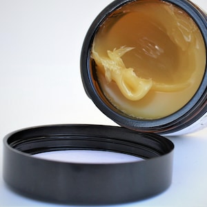 May include: A partially filled jar of a yellow-coloured balm or cream with a black lid. The jar is open and the lid is sitting next to it.