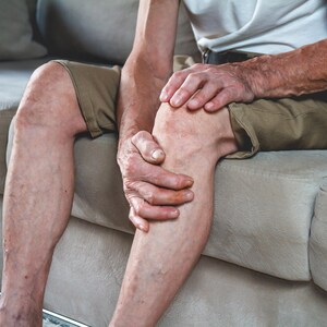 May include: A person sitting on a couch, holding their knee with both hands. They are wearing khaki shorts and a white t-shirt.