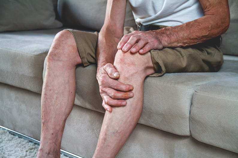May include: A person sitting on a couch, holding their knee with both hands. They are wearing khaki shorts and a white t-shirt.