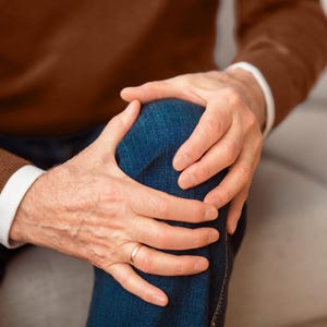 May include: A person is holding their knee with both hands, showing signs of pain or discomfort. They are wearing a brown sweater and blue jeans.