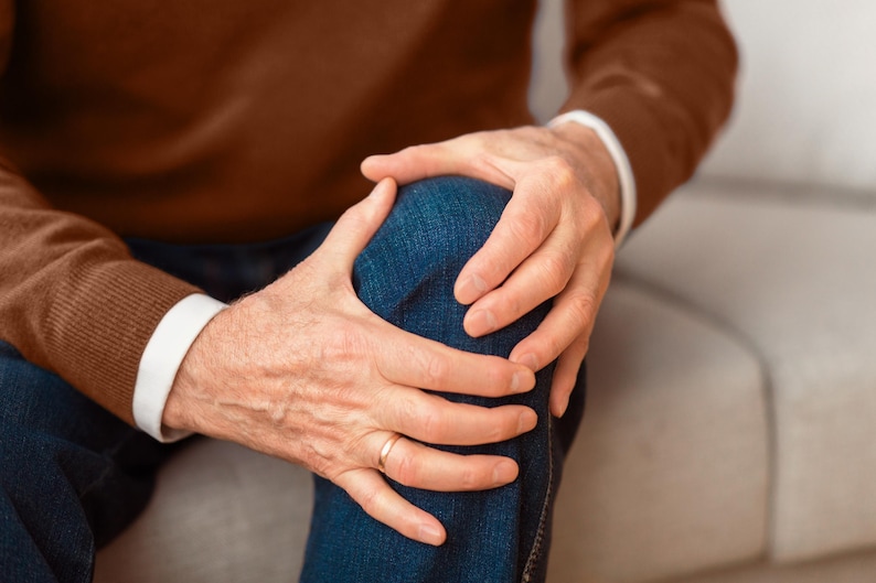 May include: A person is holding their knee with both hands, showing signs of pain or discomfort. They are wearing a brown sweater and blue jeans.