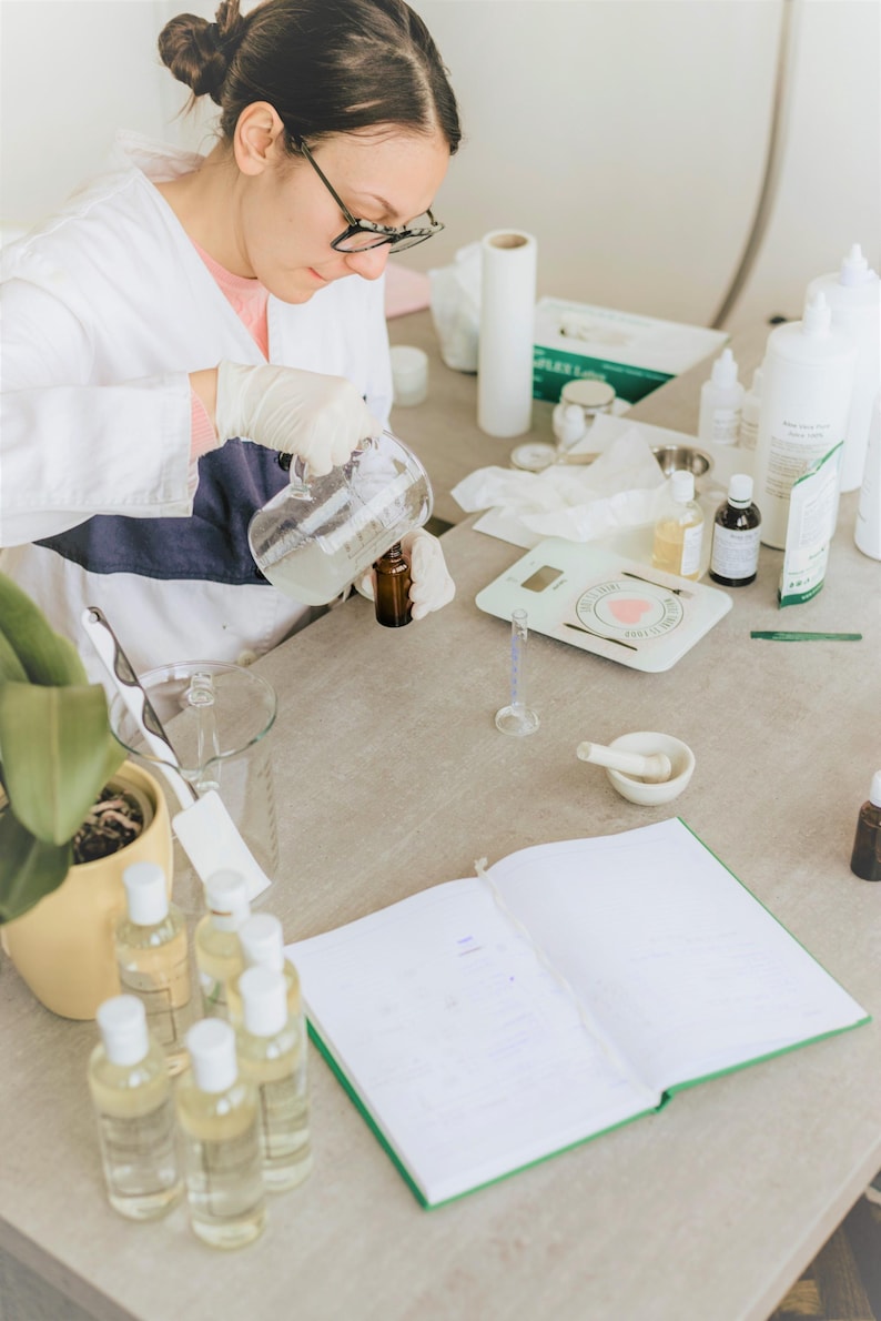 May include: A person wearing a white lab coat and gloves pours liquid from a glass beaker into a small bottle. The person is working at a table with a notebook, a scale, and other bottles and containers.