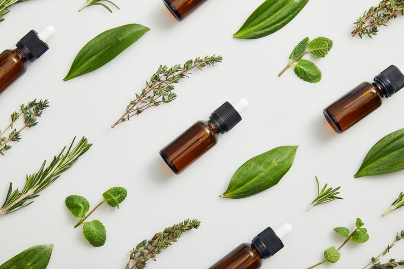 May include: A flat lay of essential oil bottles with droppers and various green herbs on a white background. The herbs include rosemary, thyme, and mint.