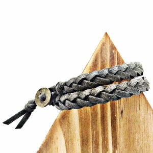 May include: A braided dark gray leather bracelet featuring a circular antique brass-toned metal bead closure. The bracelet is constructed from multiple strands of woven leather cord that wrap around to form a layered appearance with two loose ends.